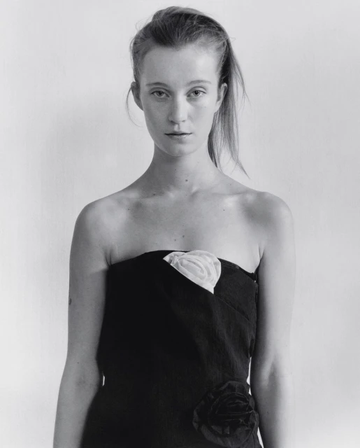 Monochrome portrait of a woman with long hair, wearing a strapless black dress with a white floral detail, against a white background.