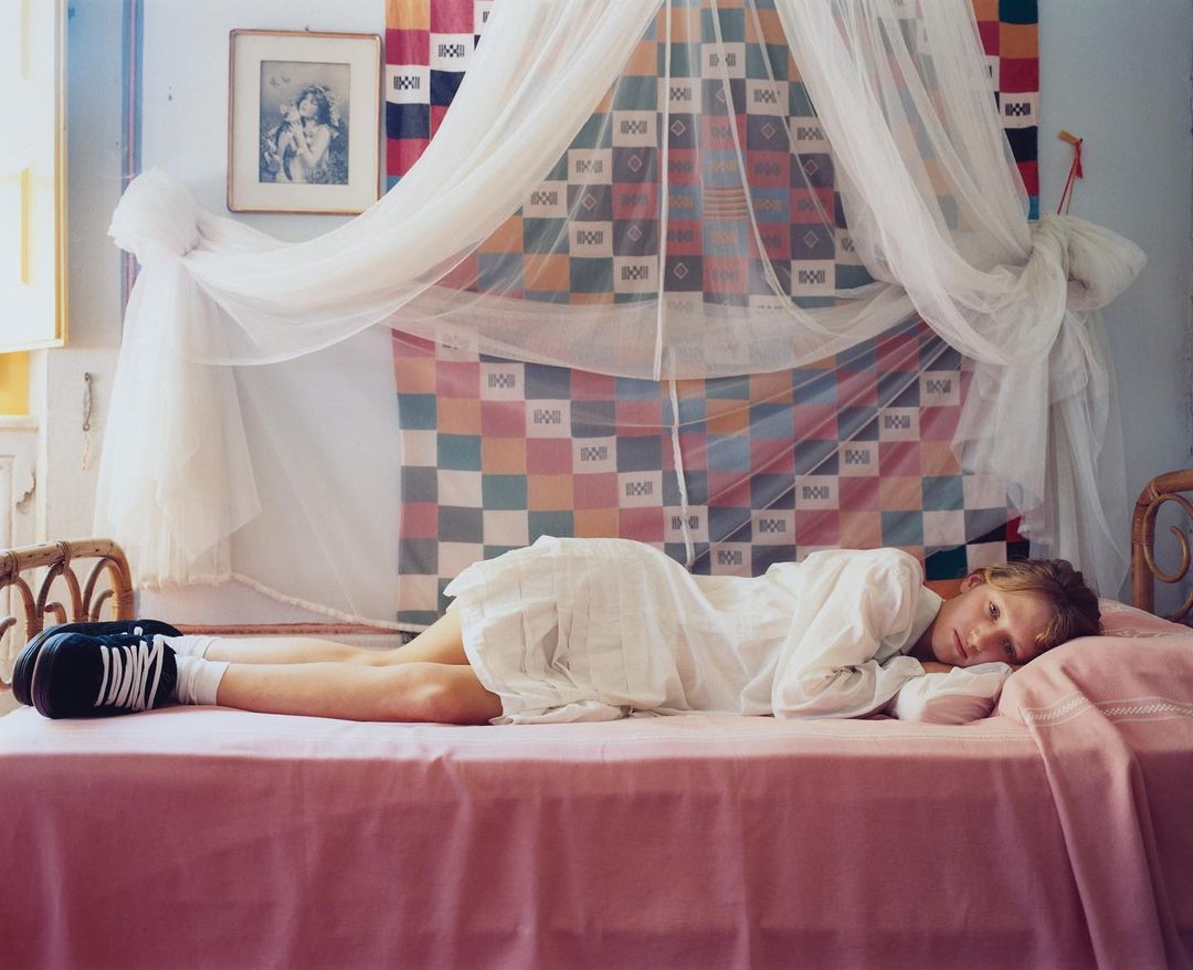 Young person with fair skin in white dress and sneakers lying on a pink bed with a mosquito net in a bright room.