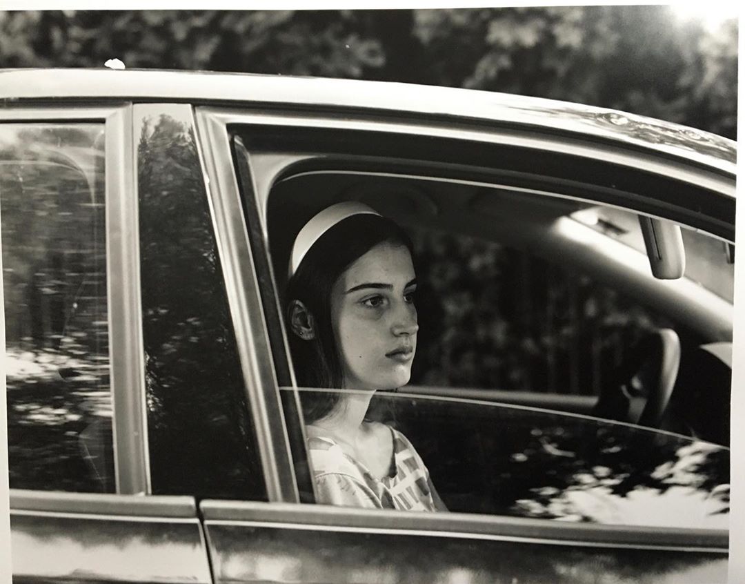 Monochrome portrait of a young woman with a headband sitting inside a car, looking out the window with trees in the background.