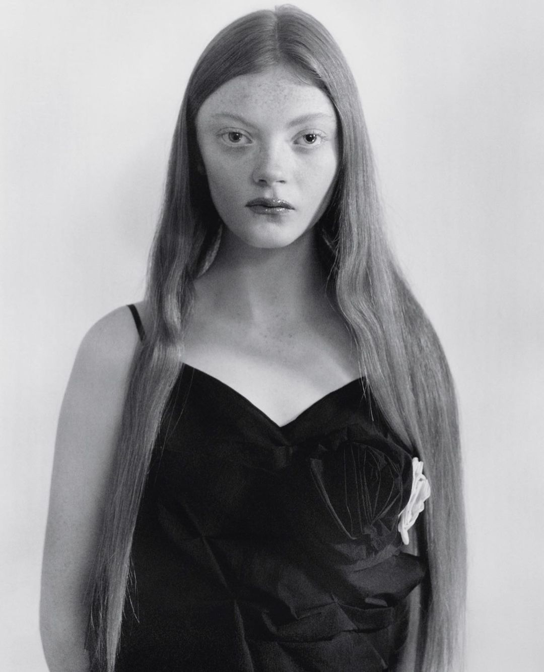 Monochrome portrait of a young woman with long hair wearing a black dress with a white floral detail, looking directly at the camera.