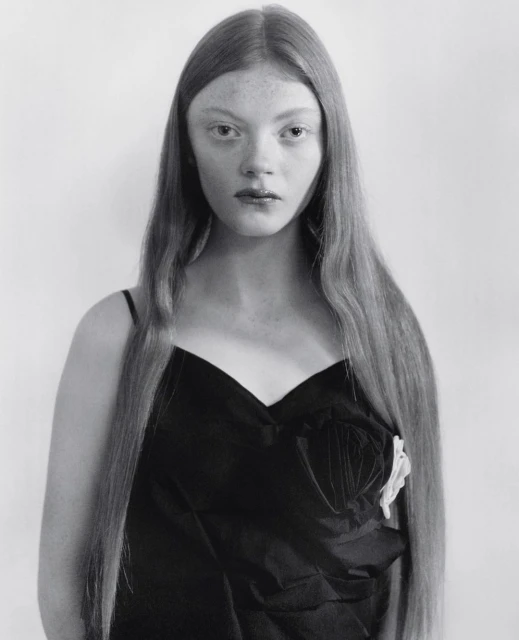 Monochrome portrait of a young woman with long hair wearing a black dress with a white floral detail, looking directly at the camera.