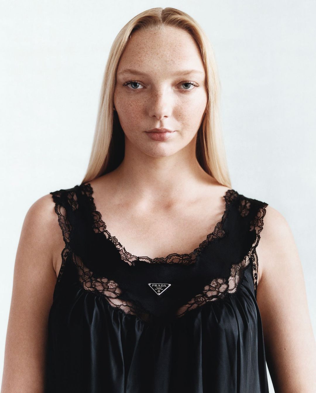 Close-up portrait of a fair-skinned young woman with blonde hair wearing a black lace-trimmed dress against a white background.