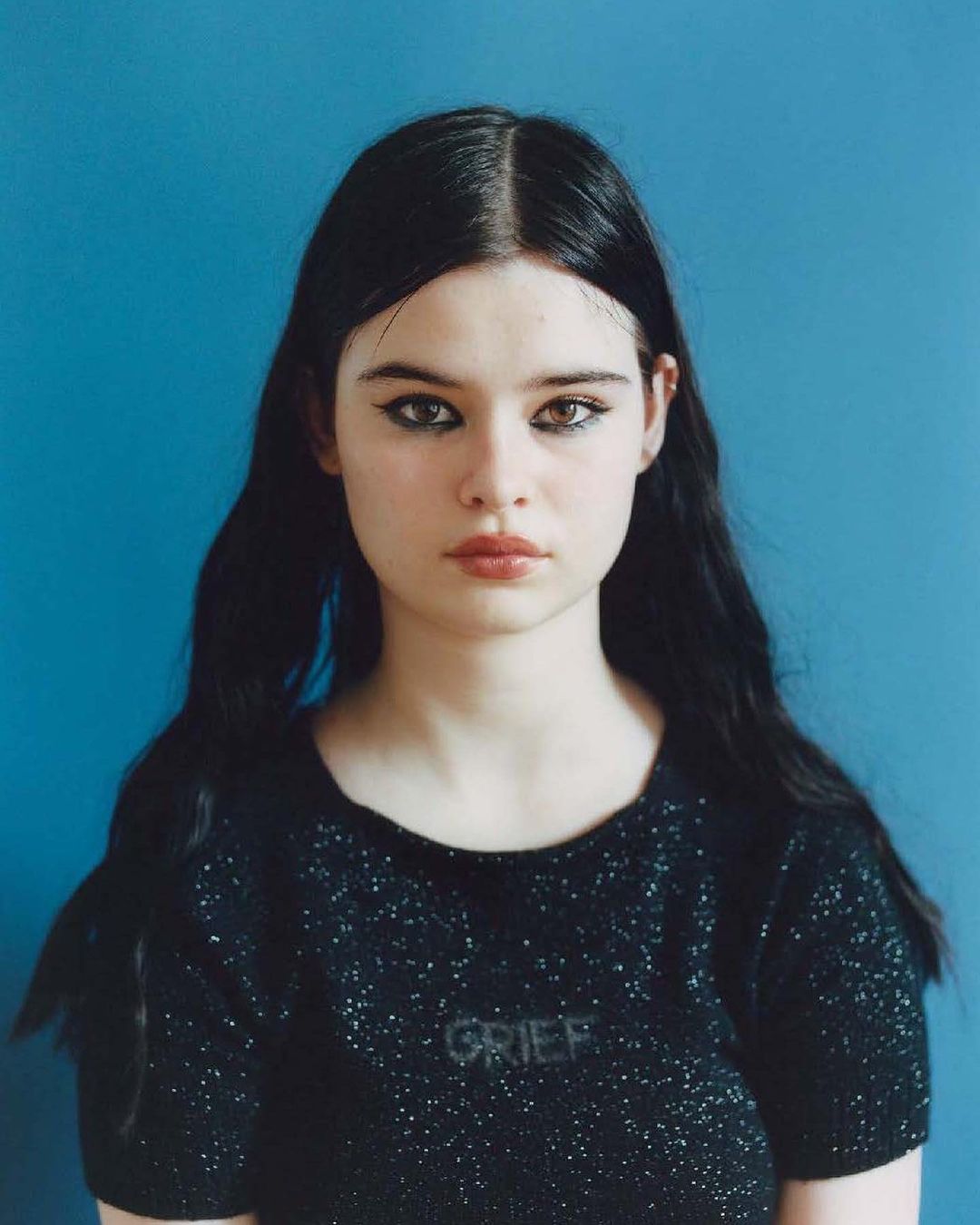 Portrait of a young woman with long black hair, wearing a black top with the word 'GRIEF' on it, against a blue background.