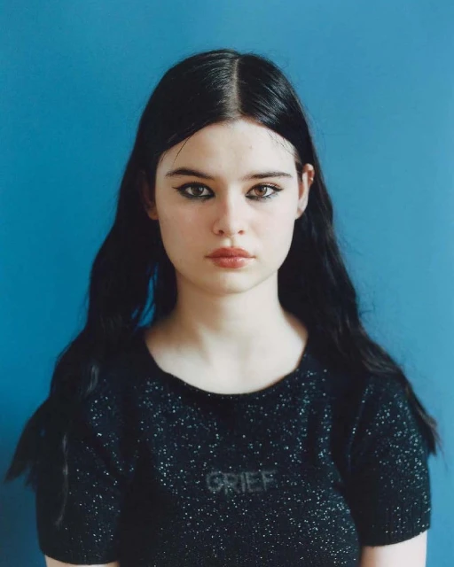 Portrait of a young woman with long black hair, wearing a black top with the word 'GRIEF' on it, against a blue background.