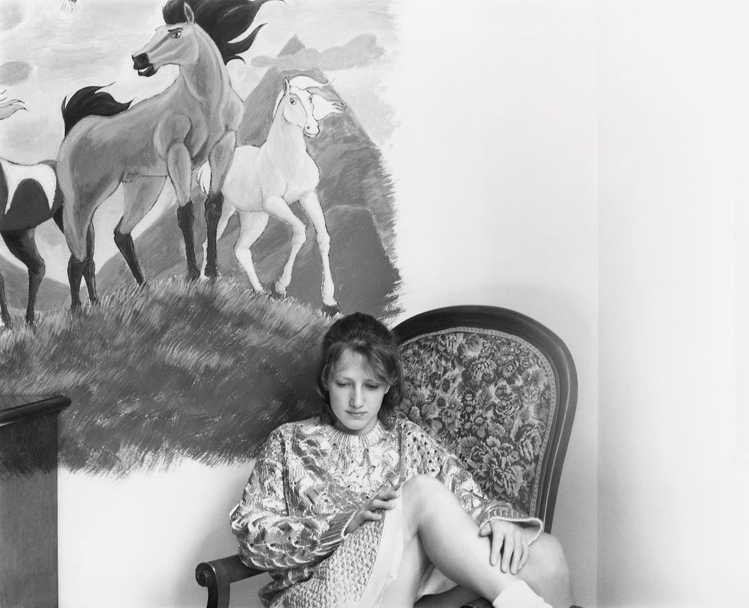 Monochrome portrait of a woman seated in a floral armchair, set against a backdrop of a wall painting featuring horses and mountains.