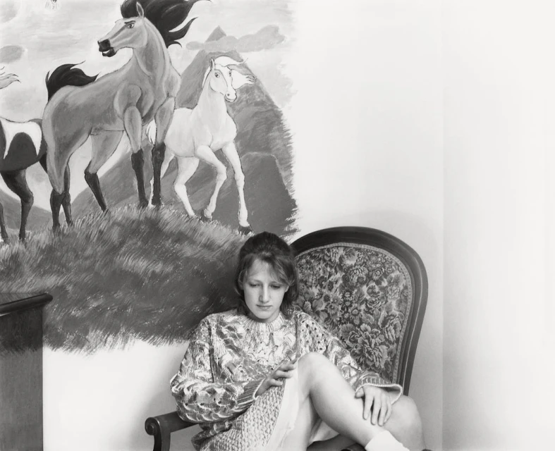 Monochrome portrait of a woman seated in a floral armchair, set against a backdrop of a wall painting featuring horses and mountains.