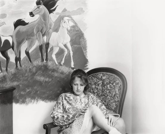 Monochrome portrait of a woman seated in a floral armchair, set against a backdrop of a wall painting featuring horses and mountains.