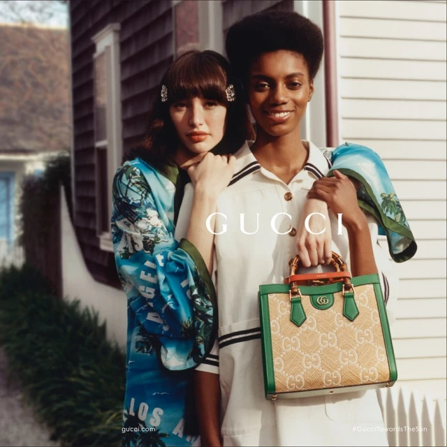 Two women pose with a Gucci handbag in a lifestyle fashion shot with a building backdrop. One woman wears a patterned robe, the other a white dress.