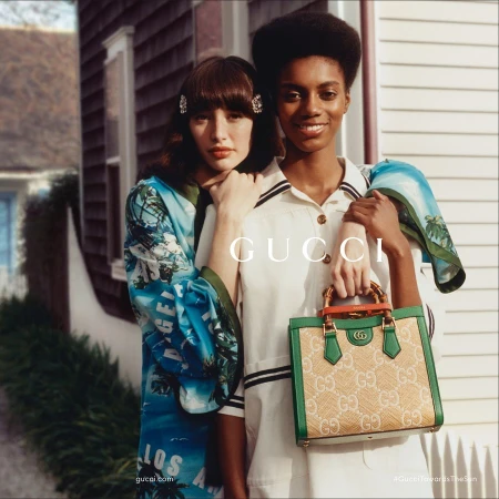 Two women pose with a Gucci handbag in a lifestyle fashion shot with a building backdrop. One woman wears a patterned robe, the other a white dress.