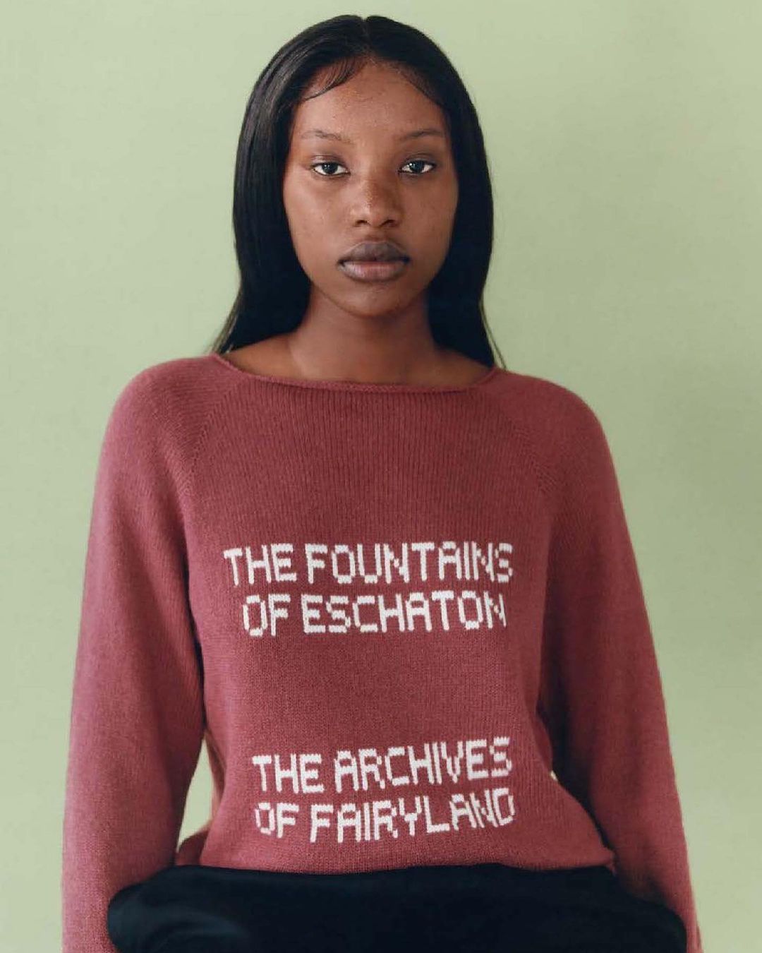 Headshot of a young adult female wearing a dark pink sweater with white block lettering on a light green studio background.