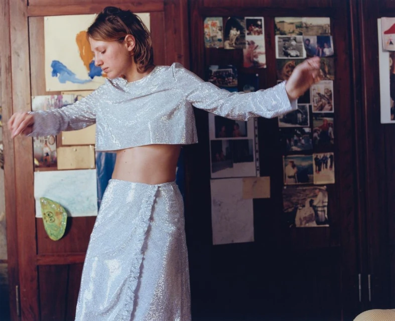 Woman in a shimmering, silvery two-piece outfit strikes a pose indoors. A wooden door with pictures serves as a backdrop, creating a casual vibe.