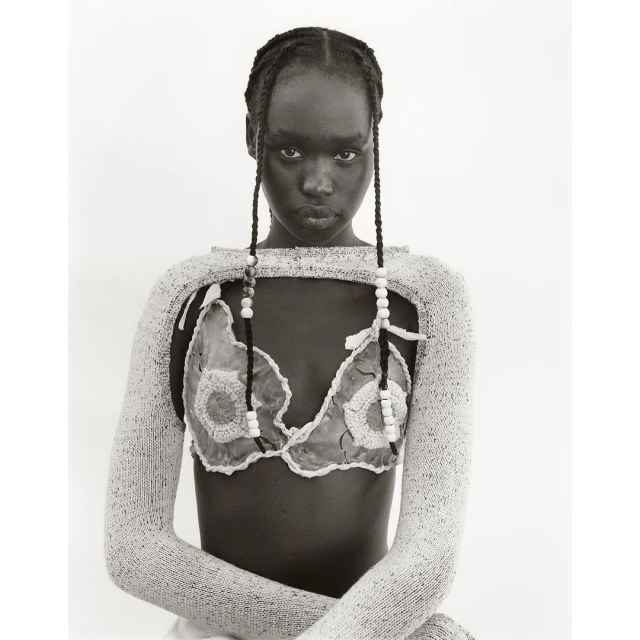 Monochromatic studio shot of a Black female model in a knit outfit with braided hair, beads, and a neutral expression against a white backdrop.