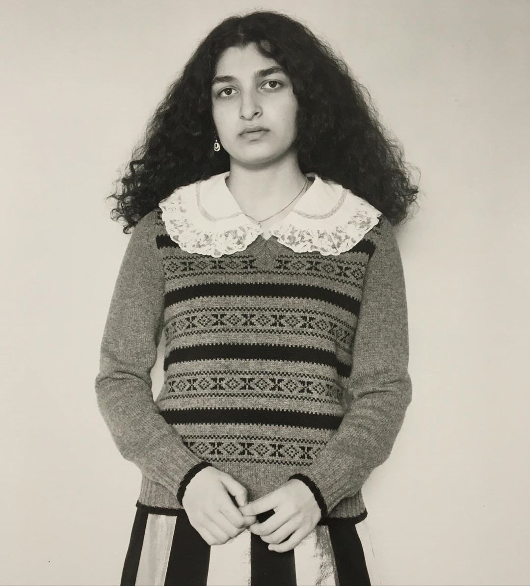 Portrait of a young adult woman with curly hair, wearing a patterned sweater with lace collar and a striped skirt, indoors.