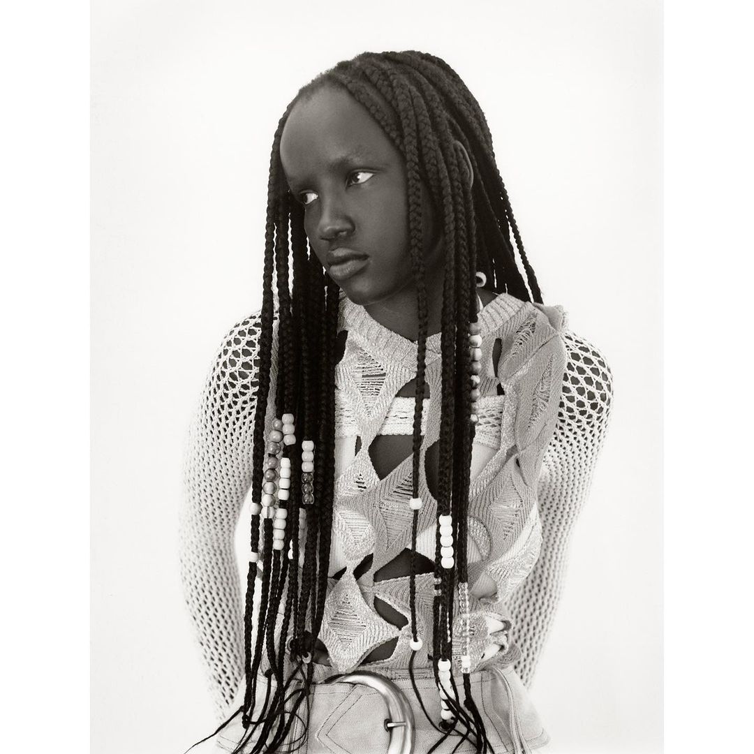 Black and white portrait of a Black woman with braided hair. She wears a light-colored, open-weave top and a belt, looking slightly away from camera.