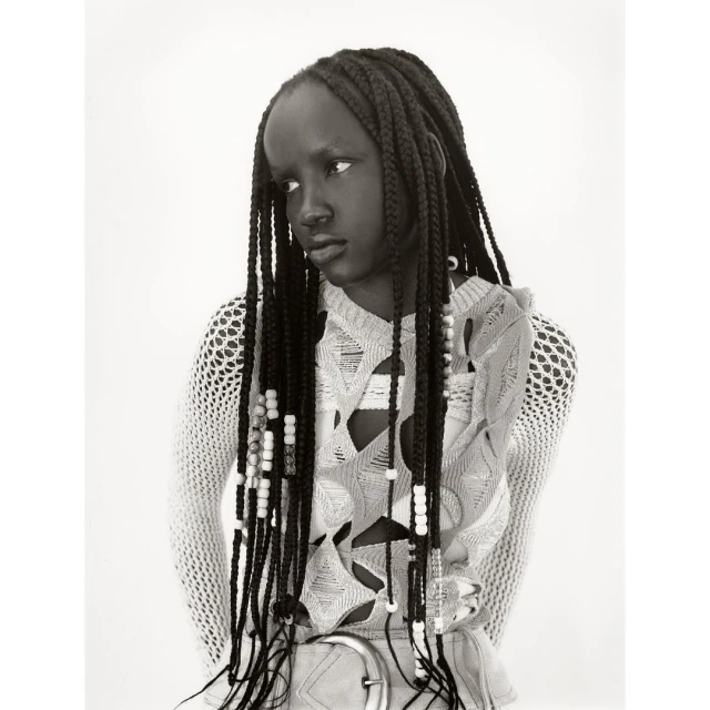 Black and white portrait of a Black woman with braided hair. She wears a light-colored, open-weave top and a belt, looking slightly away from camera.