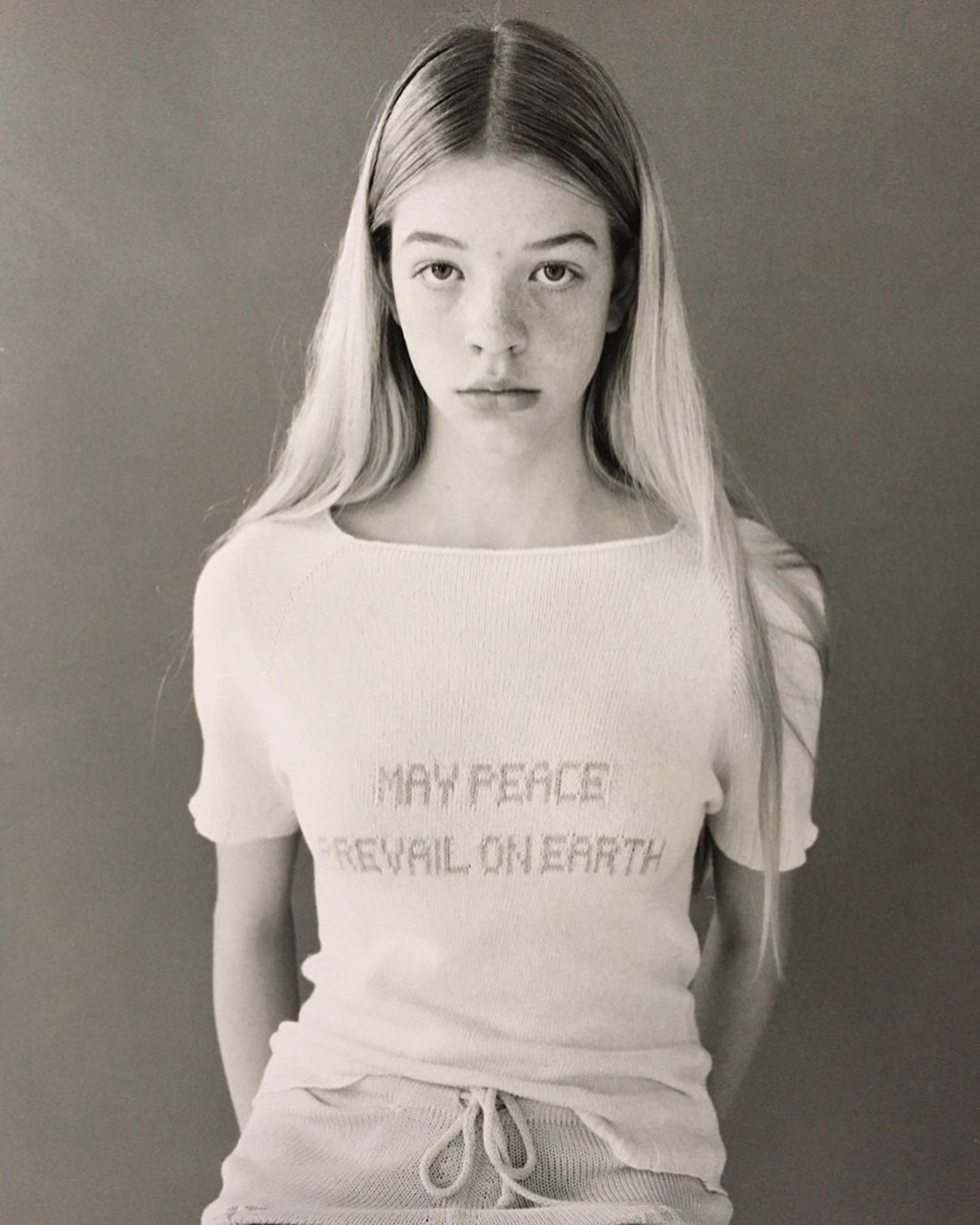 Portrait of a young woman with long blonde hair wearing a white shirt with the text, 'May Peace Prevail on Earth' on a gray background.