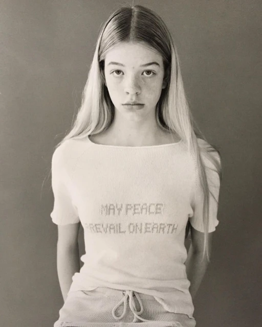 Portrait of a young woman with long blonde hair wearing a white shirt with the text, 'May Peace Prevail on Earth' on a gray background.