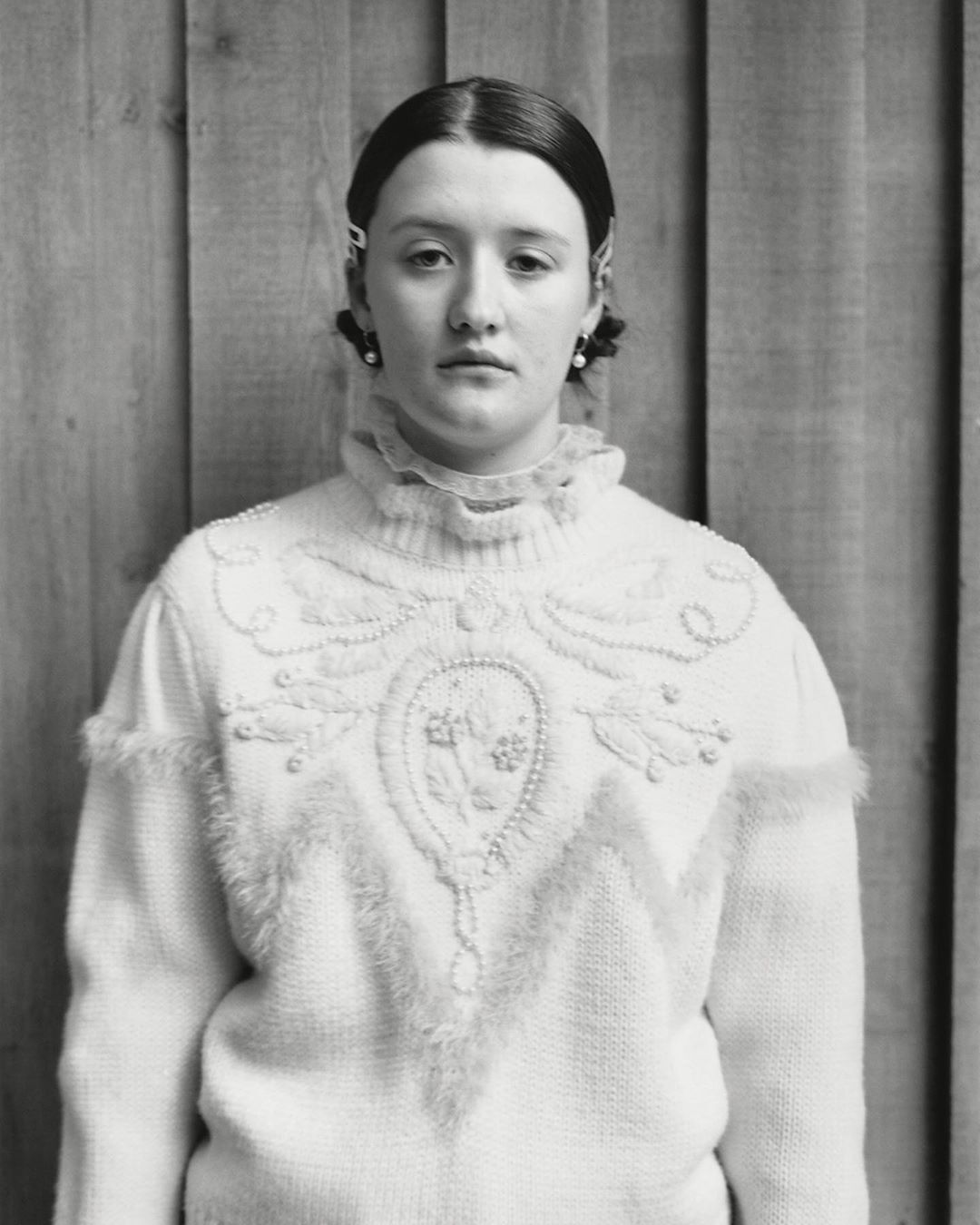 Monochrome portrait of a young woman in an embellished white sweater against a wood panel backdrop, captured in a studio setting.