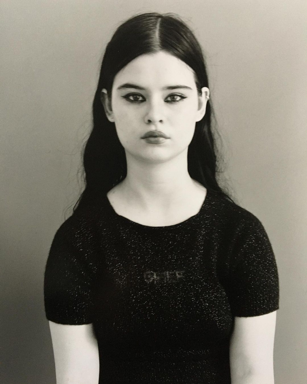 Monochrome portrait of a young woman with dark hair and heavy eye makeup, wearing a black top, against a neutral gray background.