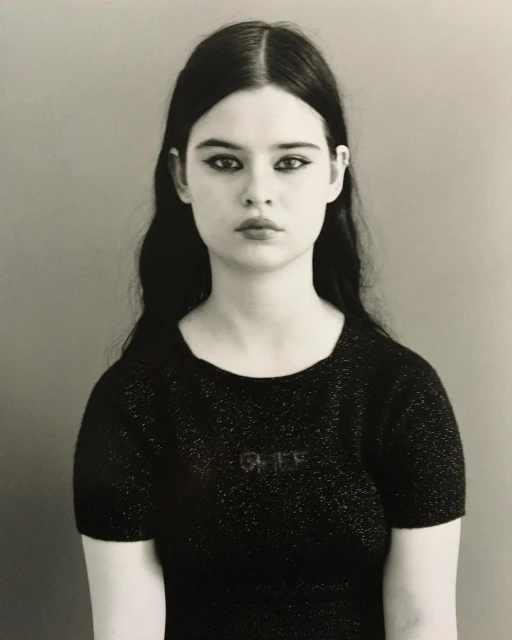 Monochrome portrait of a young woman with dark hair and heavy eye makeup, wearing a black top, against a neutral gray background.