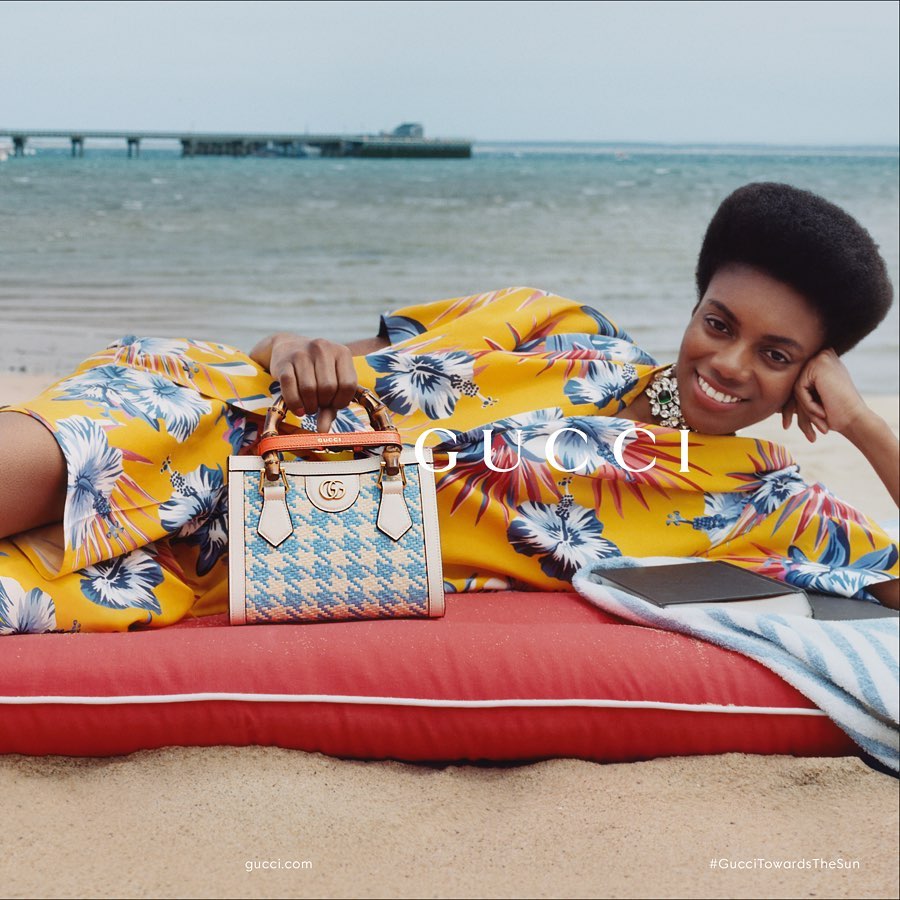 Woman in floral dress poses on beach with Gucci purse. Beach, ocean, pier, and sand provide a backdrop for fashion advertisement.