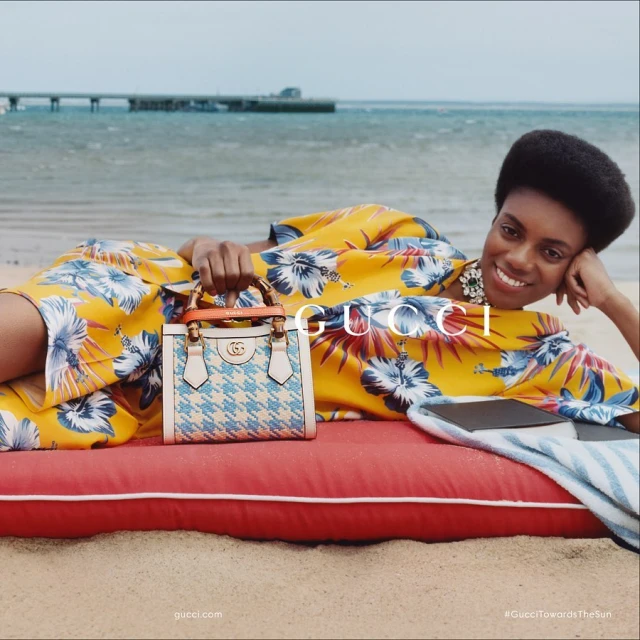 Woman in floral dress poses on beach with Gucci purse. Beach, ocean, pier, and sand provide a backdrop for fashion advertisement.