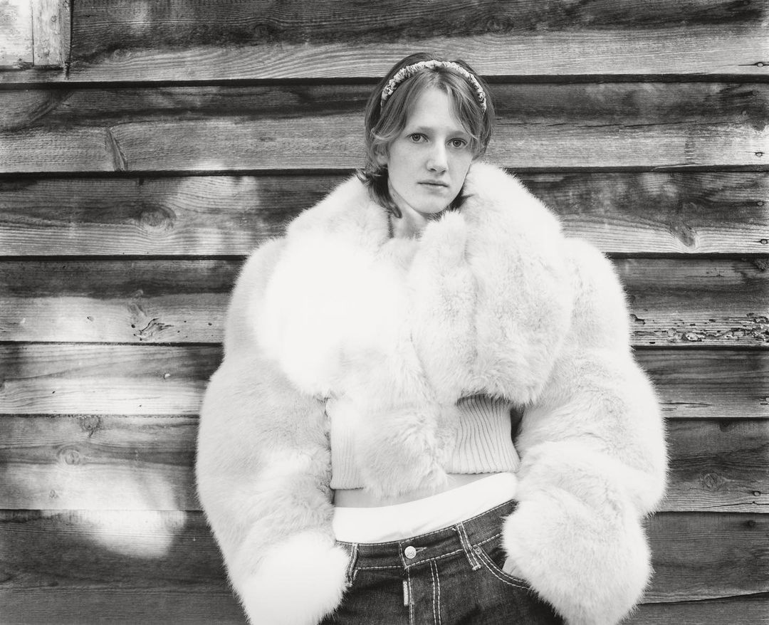 Monochrome portrait of a young woman in a fur coat against a wood plank wall, with a headband and dark jeans.