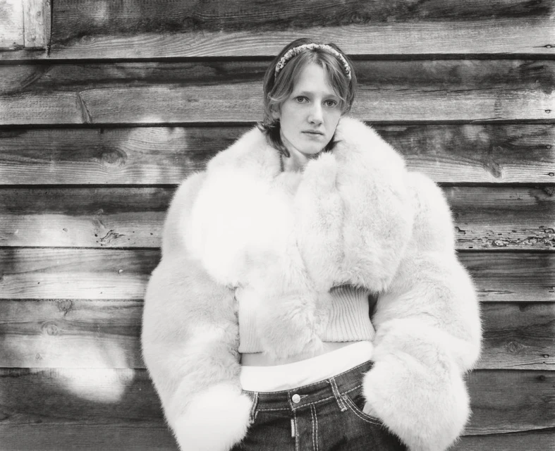 Monochrome portrait of a young woman in a fur coat against a wood plank wall, with a headband and dark jeans.