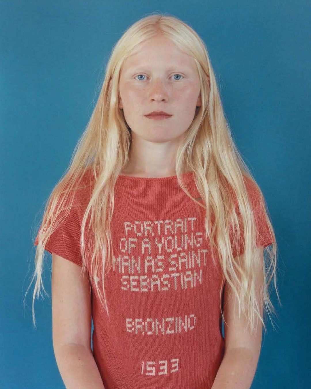 Young woman with albinism wearing a graphic knit top with text on a blue seamless background in a studio portrait, medium shot.