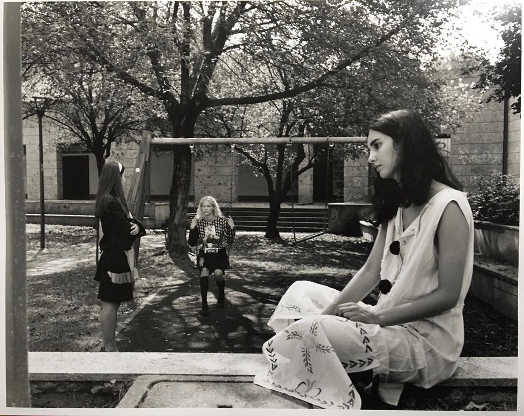 Three women in retro-style clothing pose in a black and white outdoor photo featuring mature trees and visible architecture behind them.