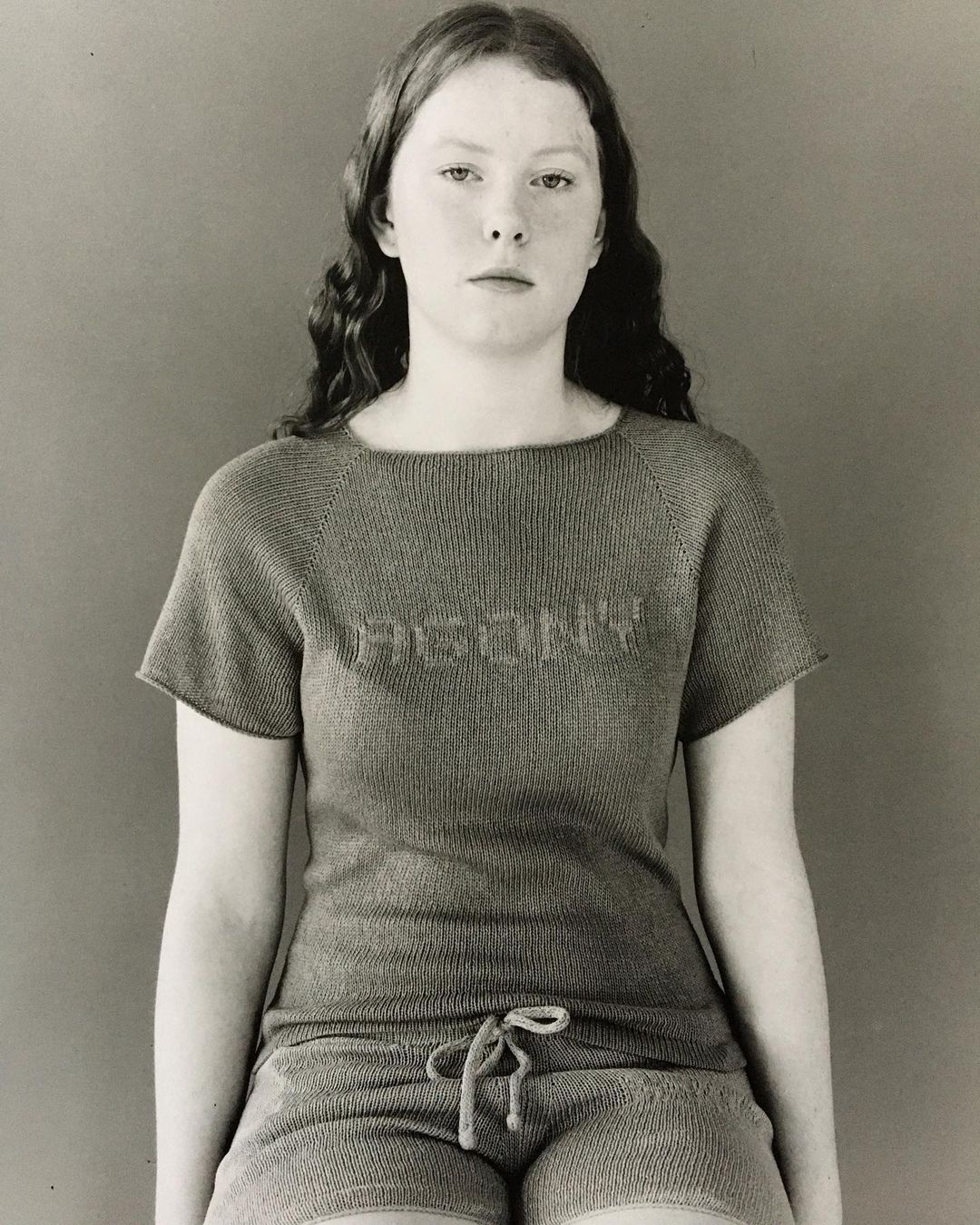 Young adult female with wavy hair wearing a knit t-shirt and shorts. Sitting indoors against a plain background in a medium shot.