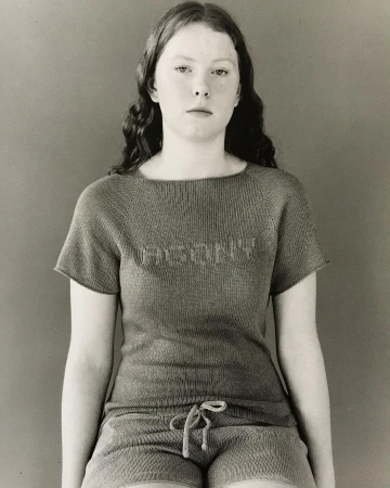 Young adult female with wavy hair wearing a knit t-shirt and shorts. Sitting indoors against a plain background in a medium shot.