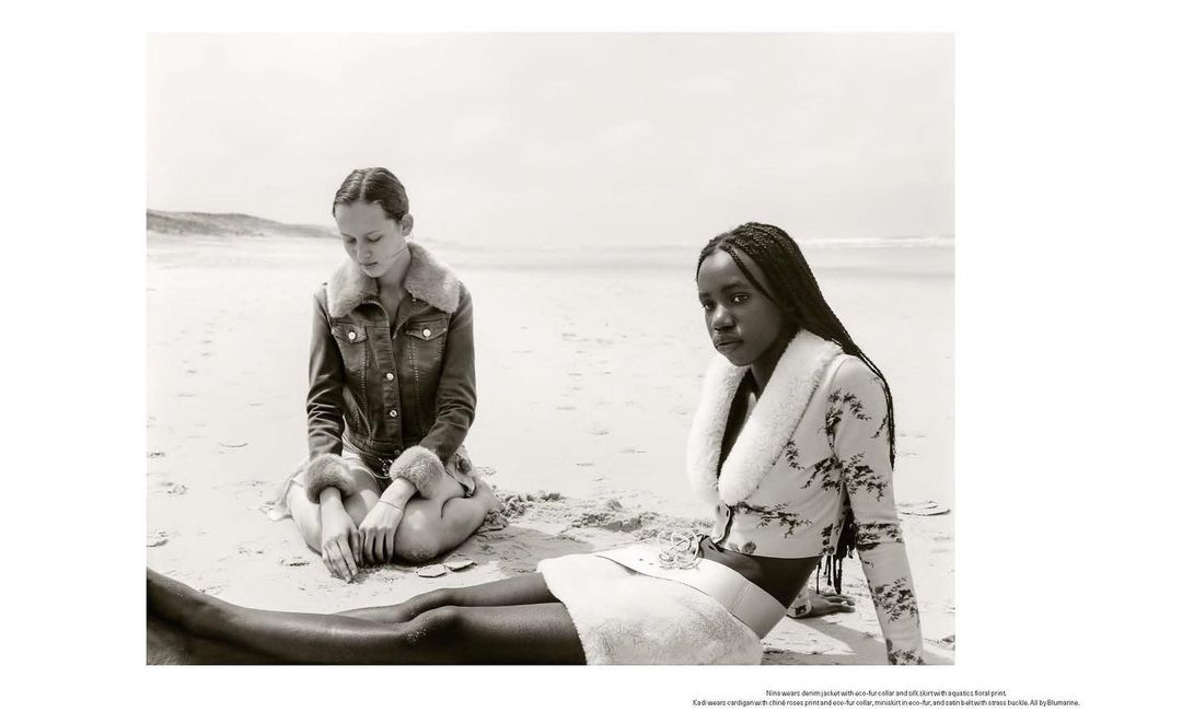 Two women sit on a sandy beach in black and white, modeling fur-trimmed jackets and skirts with the ocean and sky in the background.