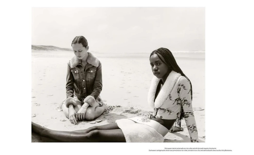 Two women sit on a sandy beach in black and white, modeling fur-trimmed jackets and skirts with the ocean and sky in the background.