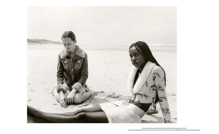 Two women sit on a sandy beach in black and white, modeling fur-trimmed jackets and skirts with the ocean and sky in the background.