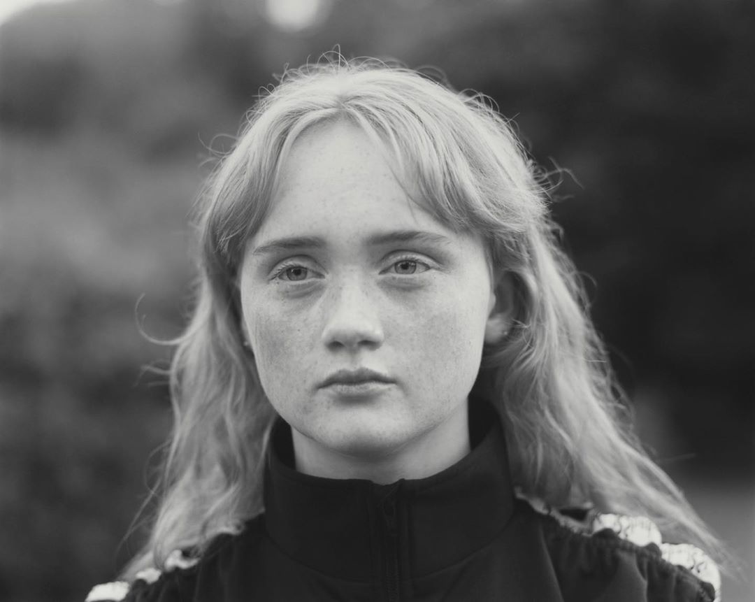 Portrait of a young woman with long wavy blonde hair, fair skin with freckles, and a dark jacket posing outdoors with a blurred background.