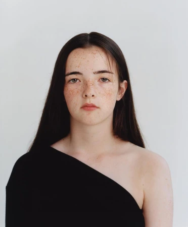 Portrait of a young woman with long dark hair and prominent freckles wearing a black off-the-shoulder top against a plain white wall