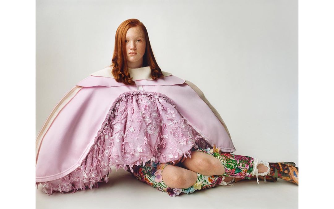 Red-haired person sitting on the floor wearing avant-garde pink fashion with floral pants in a studio with a neutral backdrop.
