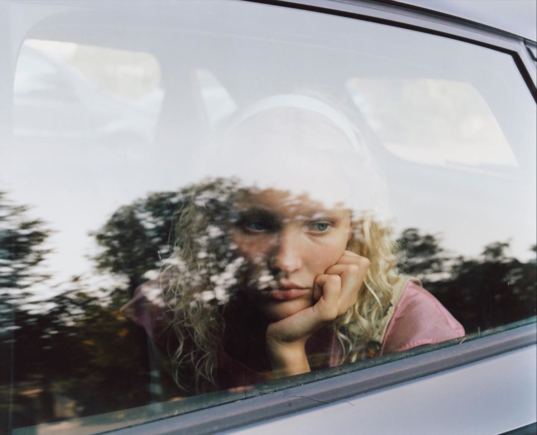 Woman with curly blond hair looks out the car window with a pensive facial expression and her head resting on her hand inside car.