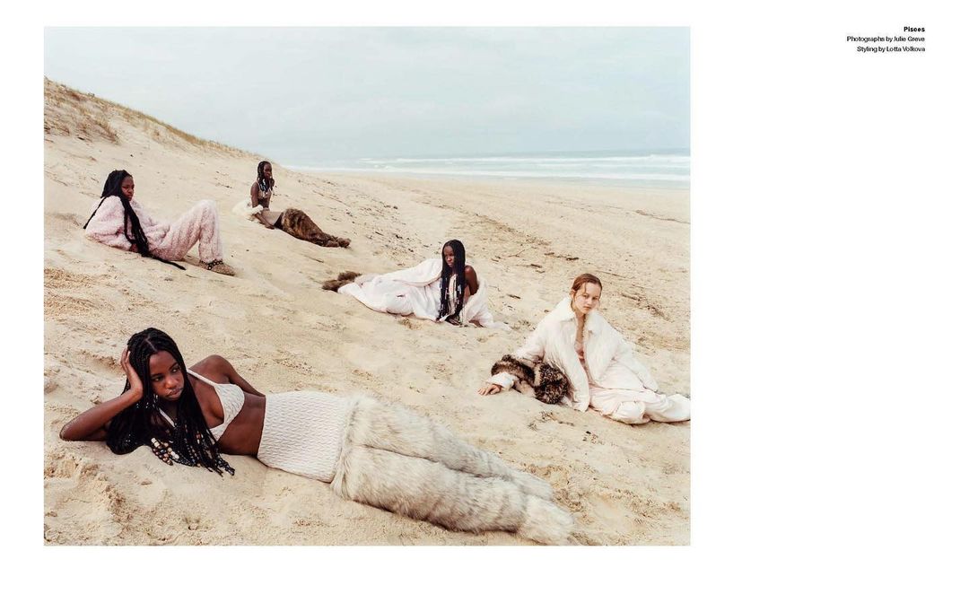 Five diverse women lounge on a sandy beach under an overcast sky. They wear various fashionable attire, including light-colored knitted clothing.