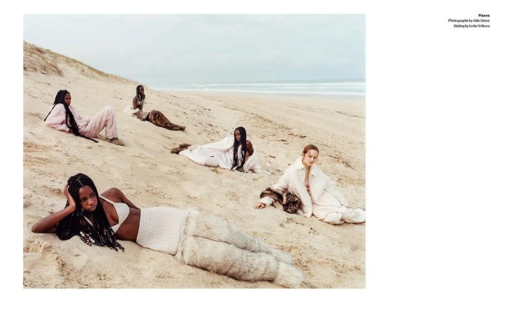 Five diverse women lounge on a sandy beach under an overcast sky. They wear various fashionable attire, including light-colored knitted clothing.