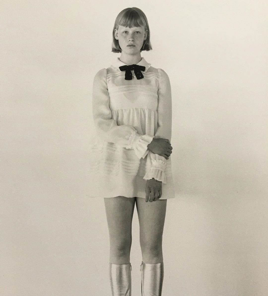 Blonde woman in white babydoll dress with black bow and silver boots, standing in front of a white background in a studio shot.