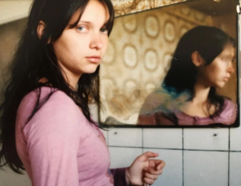 Young woman with long dark hair, wearing a pink long-sleeved shirt, looks off camera in an indoor bathroom with a mirror.