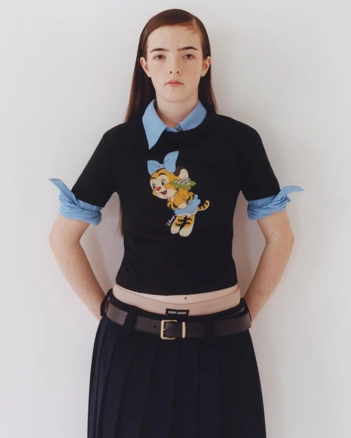 Young woman poses indoors wearing a black shirt with a cartoon tiger graphic, blue collared shirt, pleated skirt, and black belt.