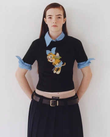 Young woman poses indoors wearing a black shirt with a cartoon tiger graphic, blue collared shirt, pleated skirt, and black belt.