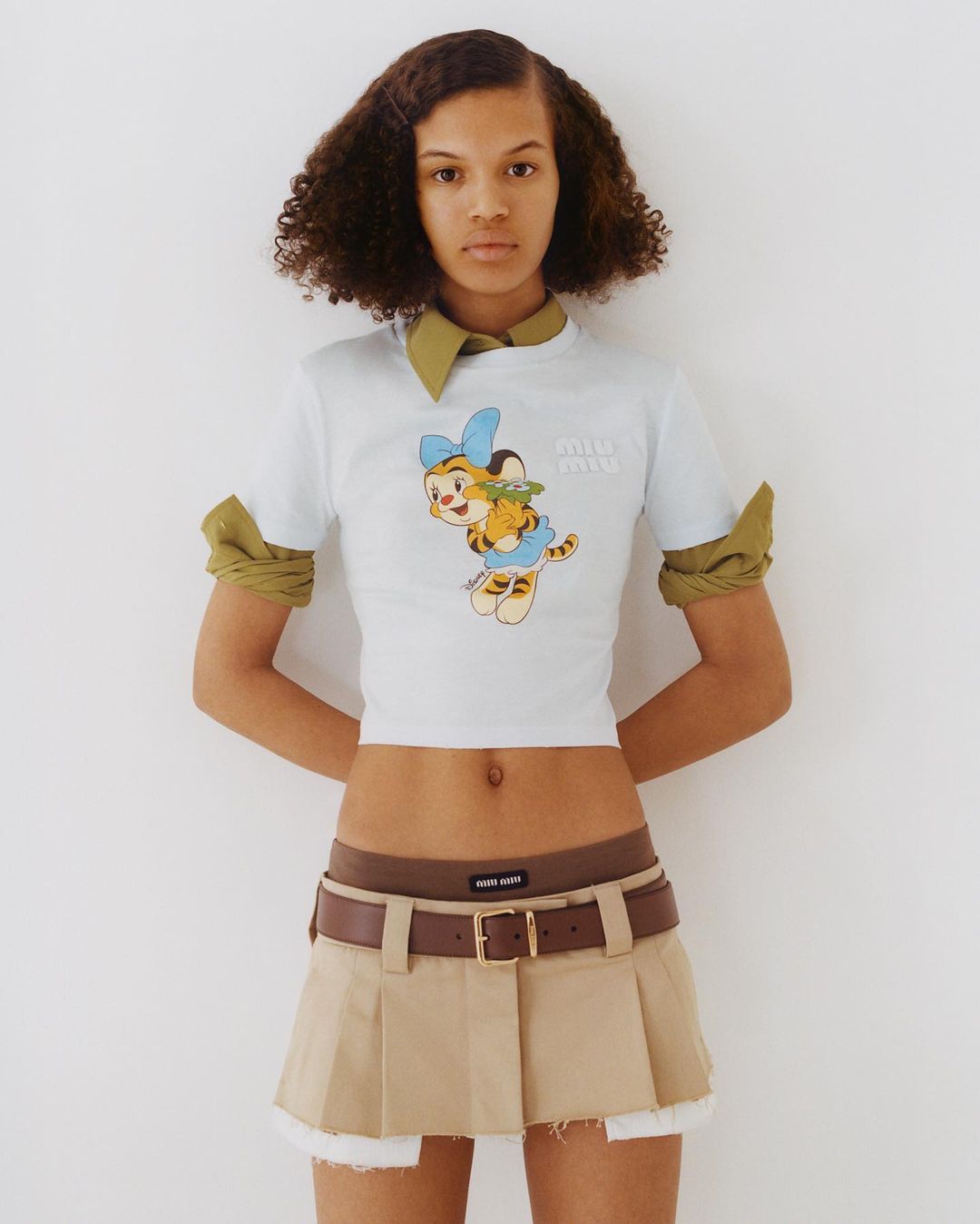 Young adult woman in a casual outfit poses indoors against a white background, showcasing a fashionable cartoon-print t-shirt and khaki skirt.