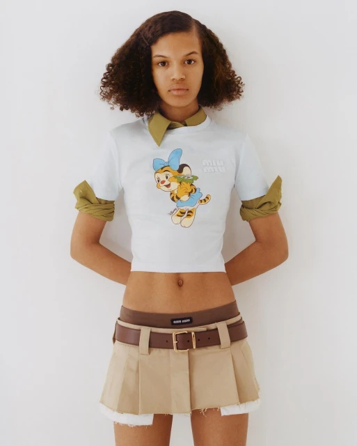 Young adult woman in a casual outfit poses indoors against a white background, showcasing a fashionable cartoon-print t-shirt and khaki skirt.