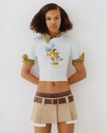 Young adult woman in a casual outfit poses indoors against a white background, showcasing a fashionable cartoon-print t-shirt and khaki skirt.