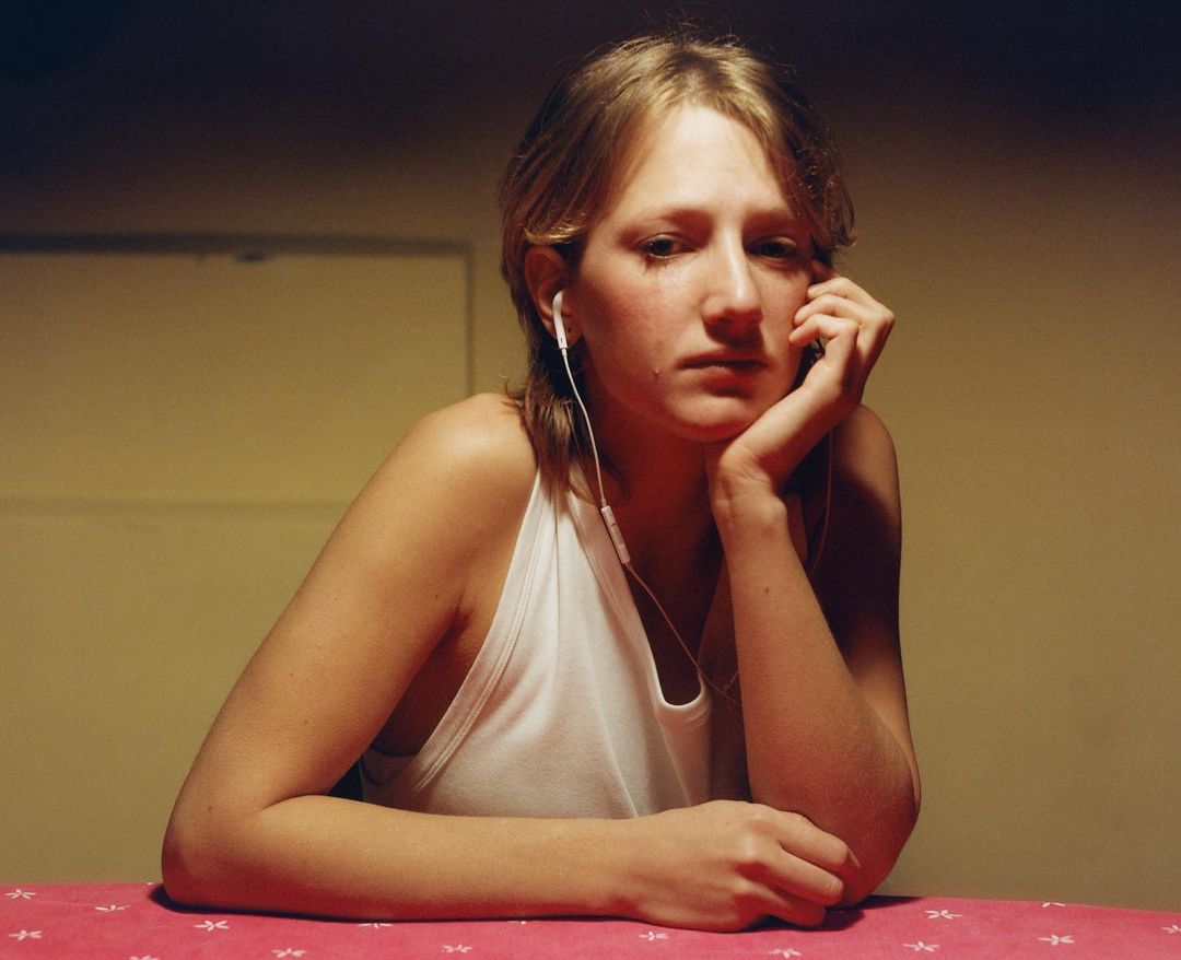 Portrait of a blond woman in a white tank top wearing headphones, sitting at a table with a pink tablecloth in an indoor setting.