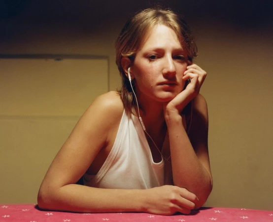 Portrait of a blond woman in a white tank top wearing headphones, sitting at a table with a pink tablecloth in an indoor setting.